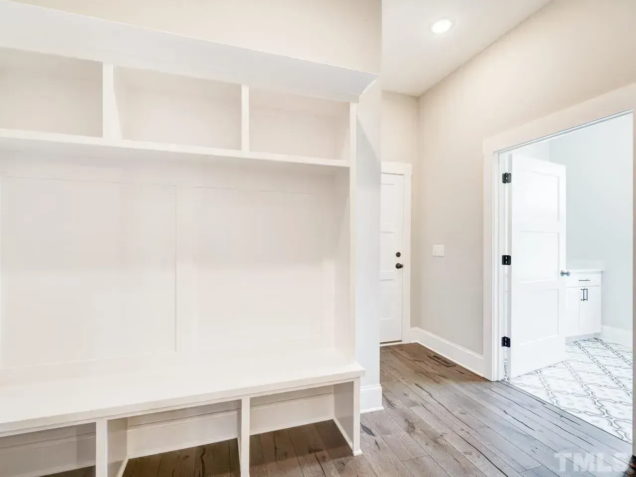 A white built-in mudroom bench with upper cubbies in a hallway with wood flooring and a doorway leading to a bathroom.