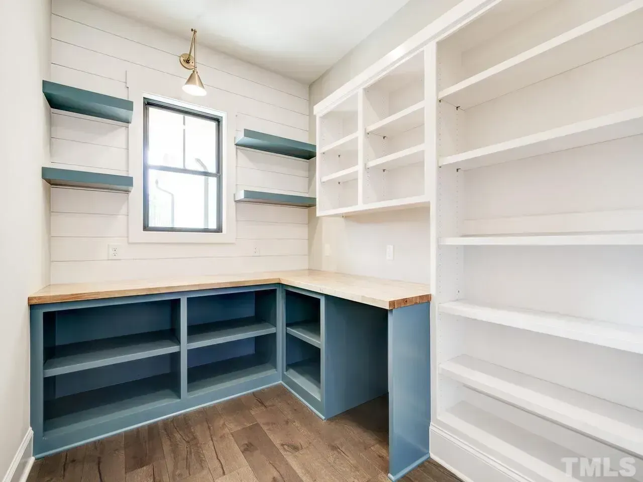 A home office or pantry featuring blue lower cabinets, a wooden countertop, white wall shelving, and a small window.
