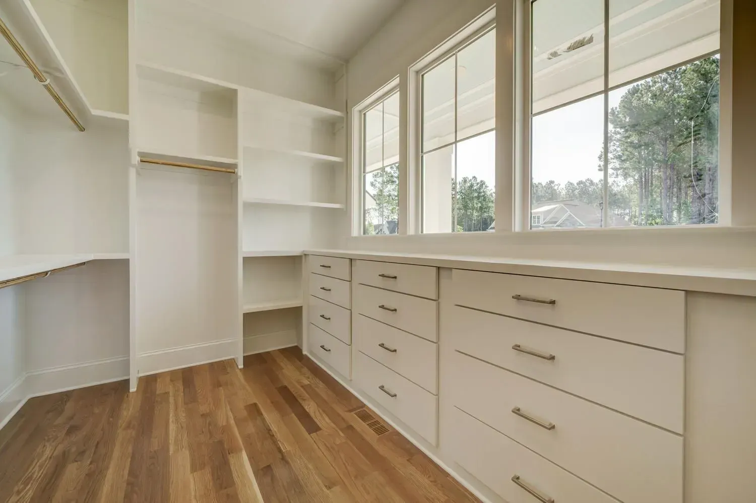 A walk-in closet featuring white wooden shelving, drawers, light hardwood floors, and large windows.