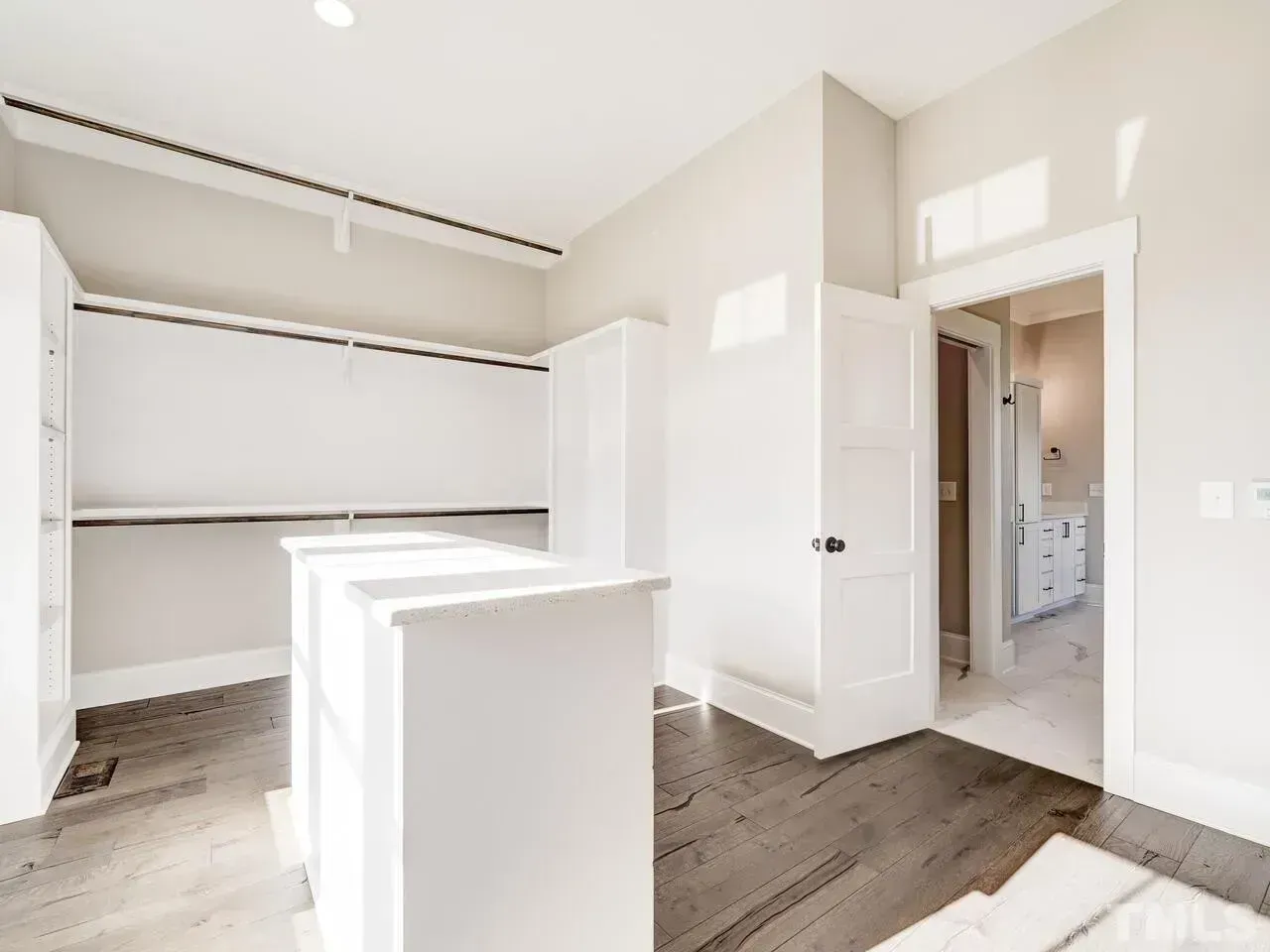 A walk-in closet featuring white shelves, a central island, and wood floors, with a doorway leading to a bathroom.