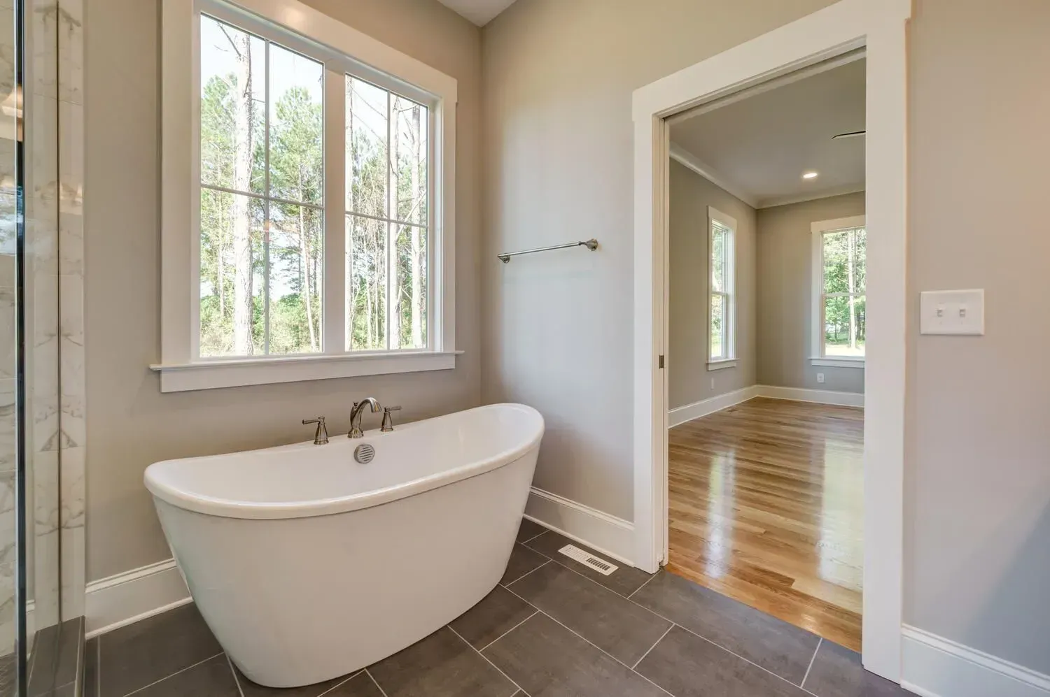 A white freestanding bathtub sits under a large window in a modern bathroom with grey tile flooring and a doorway.