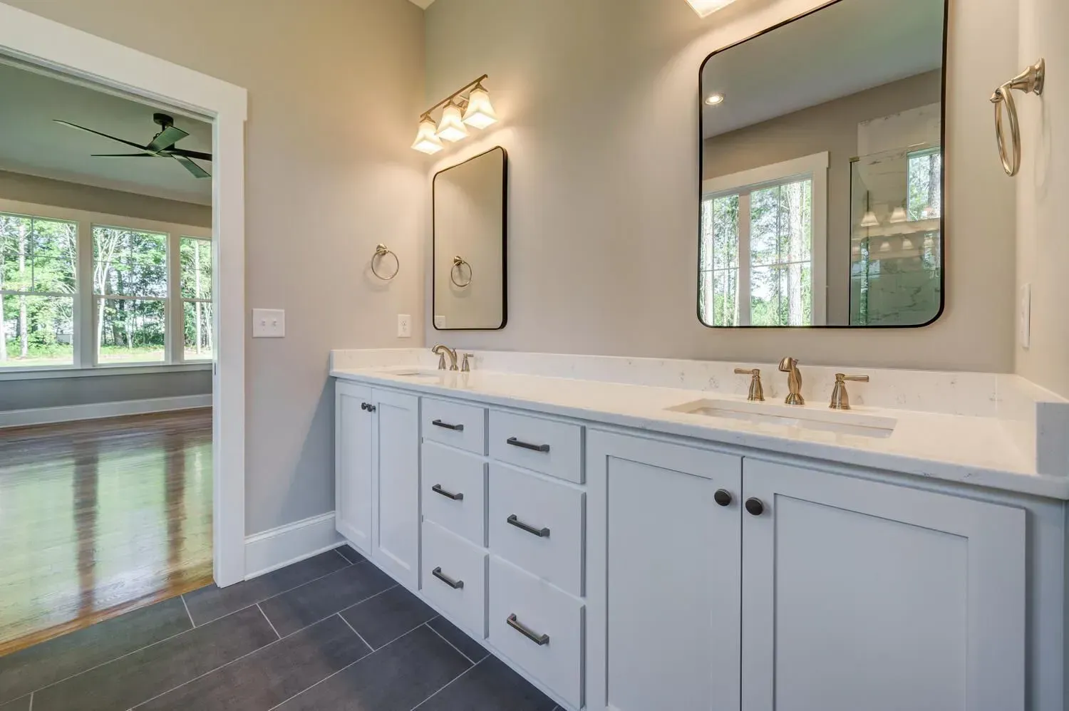 A bright bathroom featuring a white vanity with gold faucets, two mirrors, and gray tile flooring.