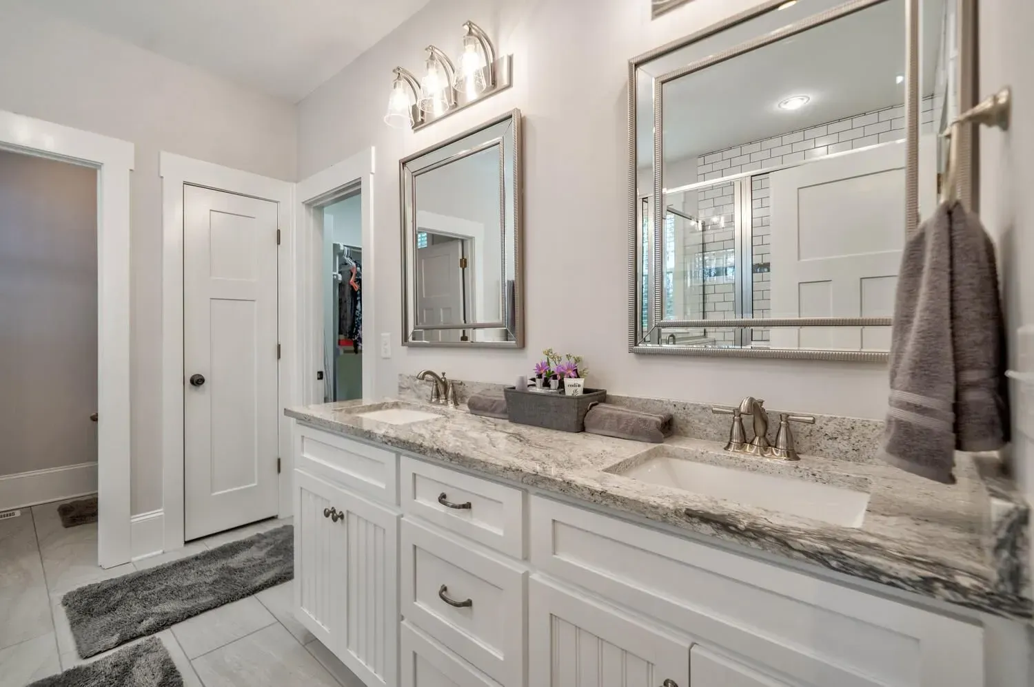 A bright, modern bathroom featuring a white vanity, dual sinks, granite countertops, two mirrors, and gray rug runners.