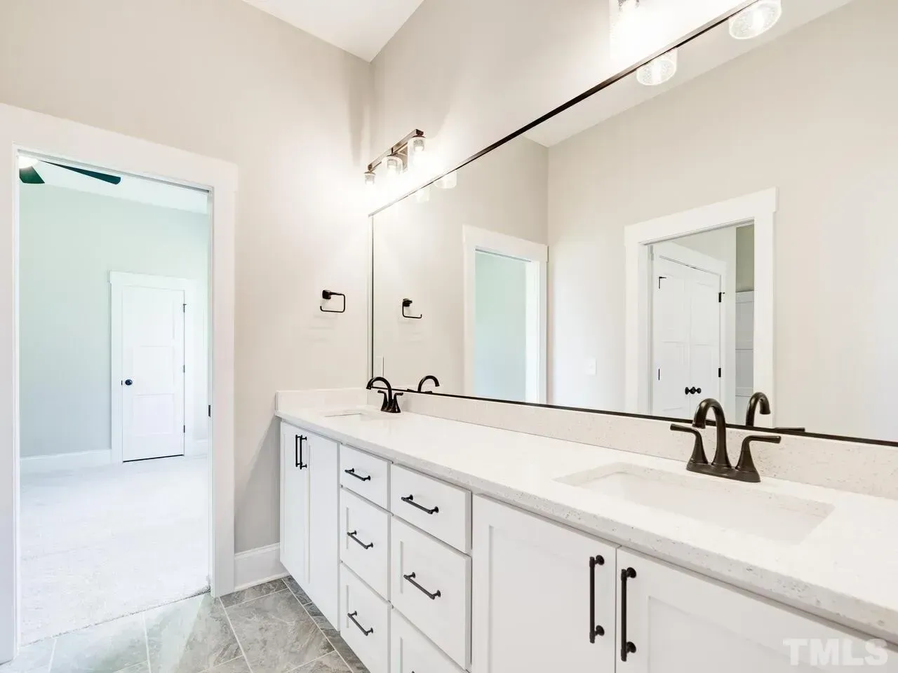 A modern bathroom with a double vanity, white cabinetry, dark hardware, and a large mirror reflecting an adjacent room.