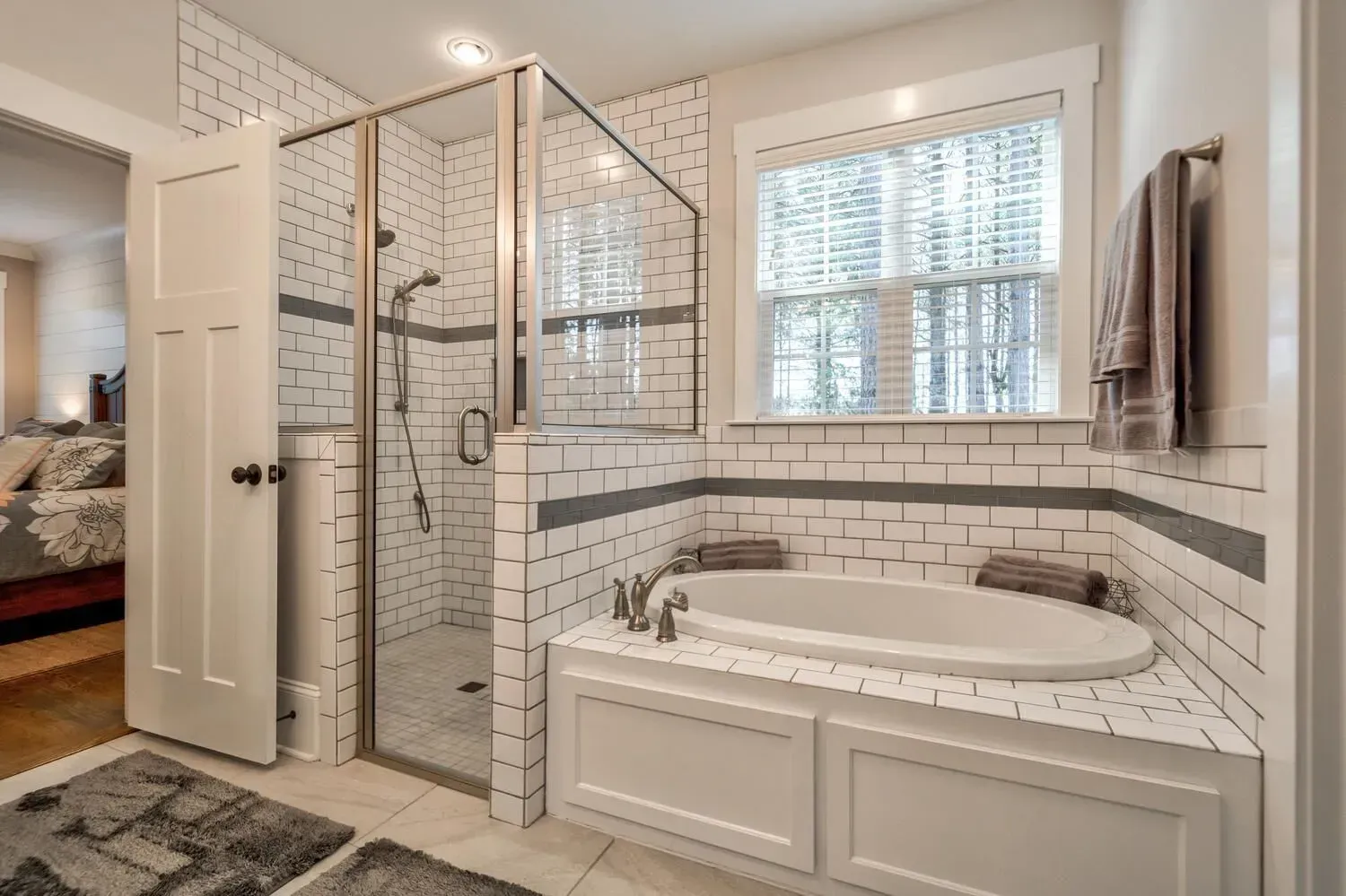 A modern bathroom featuring a white-tiled shower, a built-in soaking tub, and a window, with a view into a bedroom.