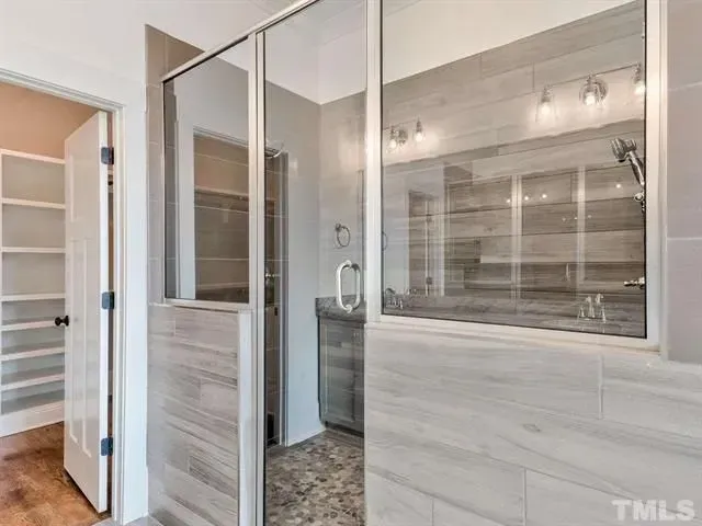 A glass-enclosed shower with light-colored tile walls and a doorway leading to a walk-in closet with white shelving.
