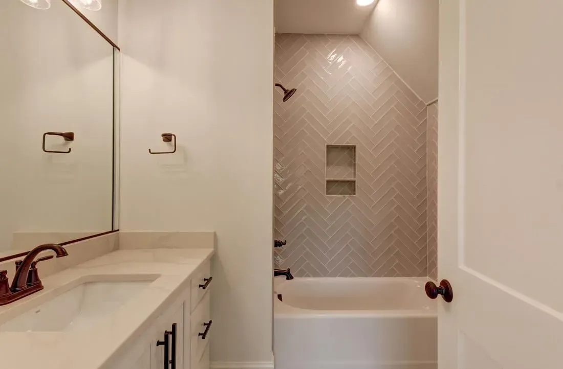 A bright bathroom featuring a white vanity with a dark faucet, a large mirror, and a tub-shower with herringbone tiles.