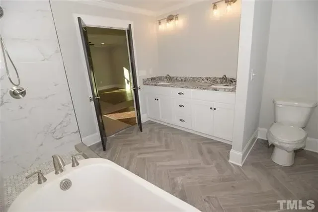 Modern bathroom with a white soaking tub, marble tiled shower, double vanity, and toilet on herringbone flooring.