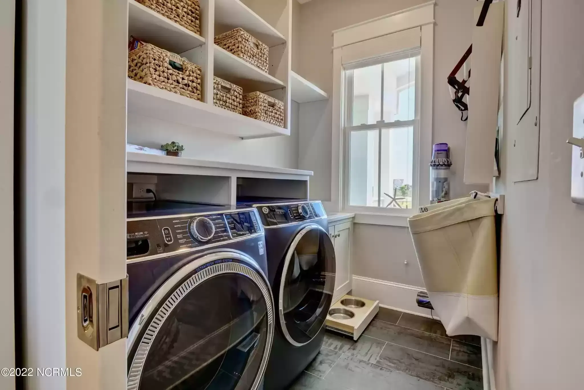 A laundry room with dark front-load machines, built-in shelving with woven baskets, and a window with natural light.