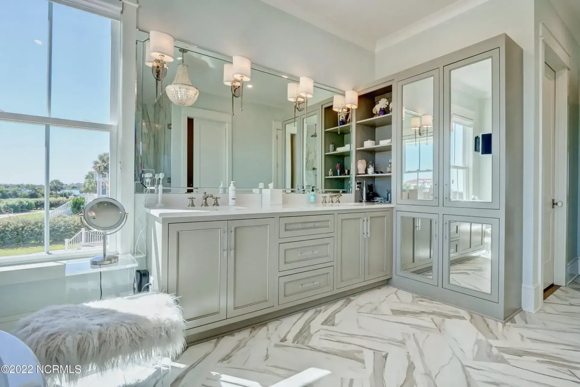 A bright bathroom featuring a light gray vanity, a large wall mirror, built-in shelving, and white marble patterned floors.