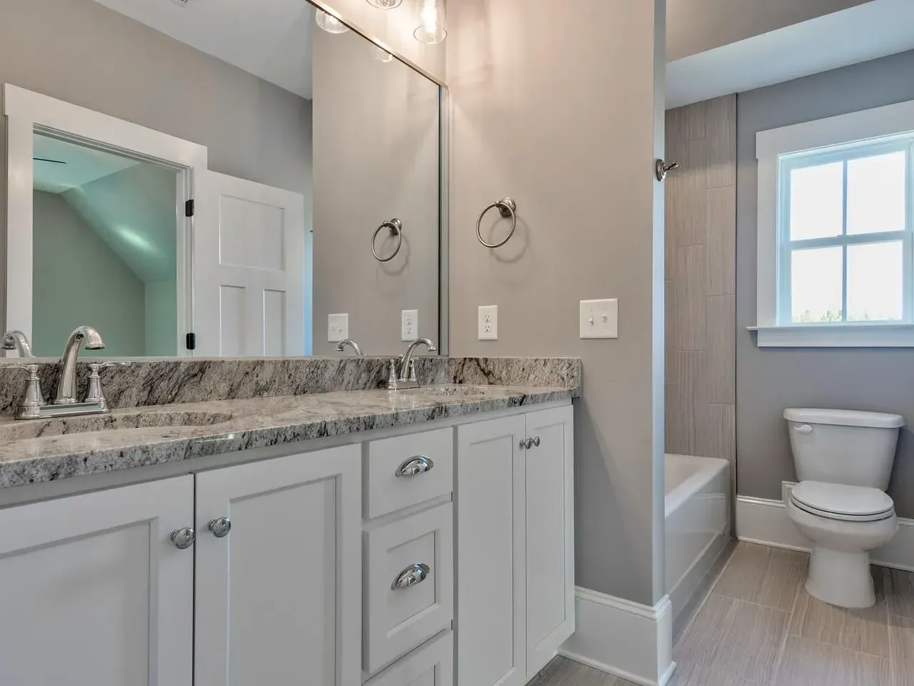 A modern bathroom with a white vanity, granite countertop, large mirror, and a toilet next to a shower alcove.