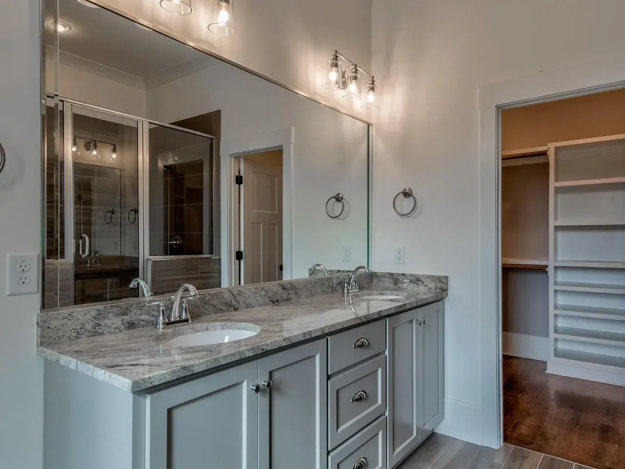 A double-sink bathroom vanity with light gray cabinets, granite countertops, and a large mirror, leading into a closet.