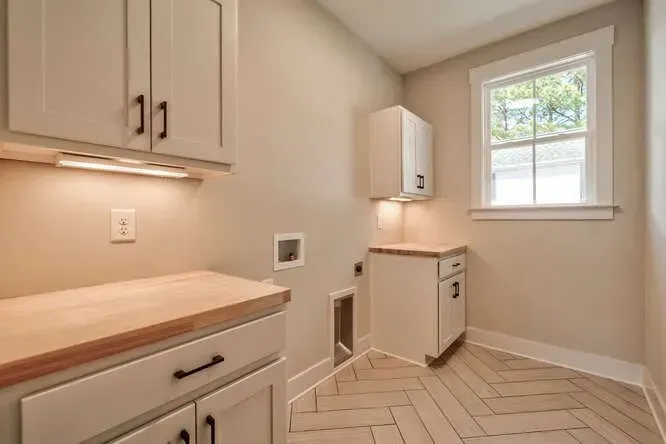 A modern laundry room with cream-colored cabinets, a butcher block countertop, a small window, and light-tiled flooring.