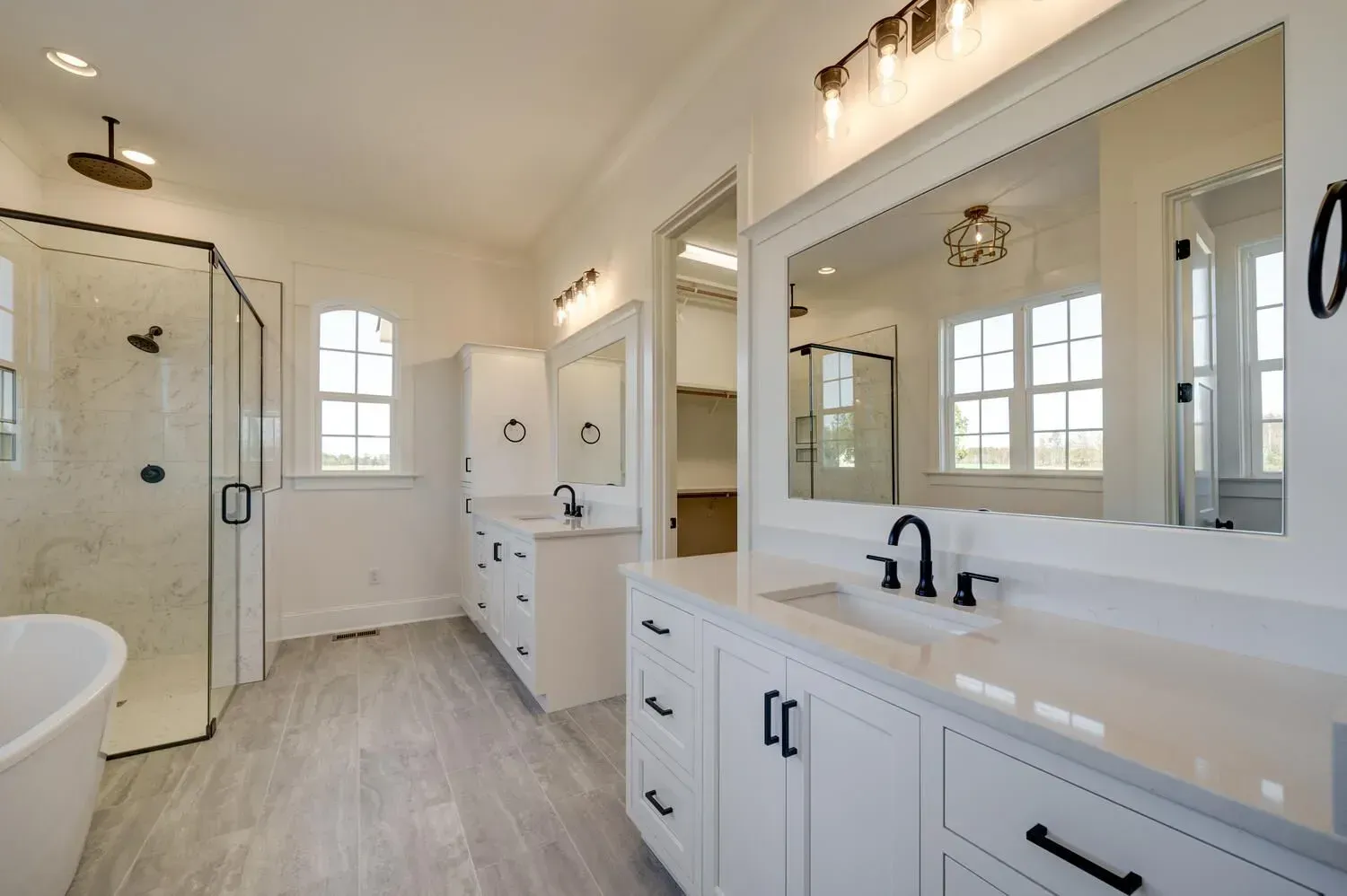 A bright, modern bathroom with a glass-enclosed shower, a white vanity with black hardware, and gray wood-look flooring.