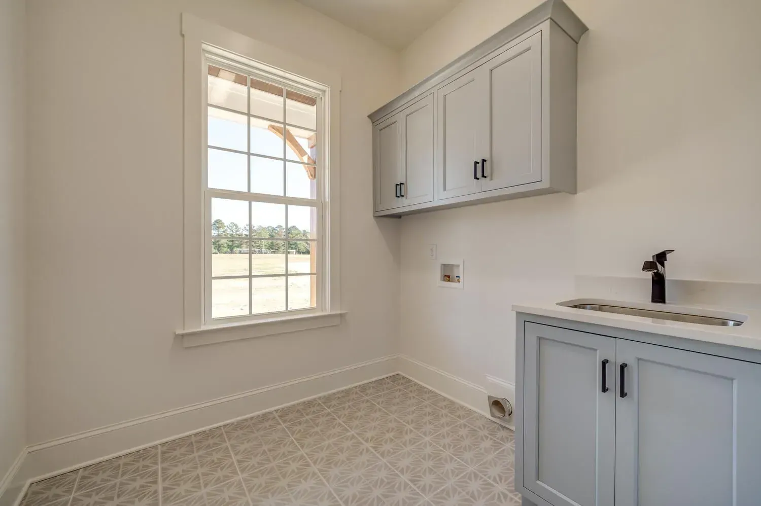 A laundry room with a window, white walls, light gray cabinets, a small sink, and a tiled floor.