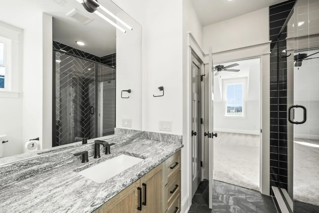 Modern bathroom featuring a granite vanity, light wood cabinets, and a glass-enclosed shower with black tiled accents.