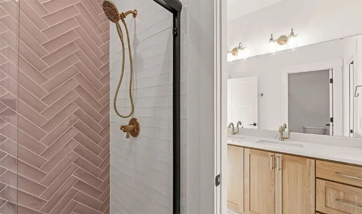 A walk-in shower with mauve herringbone tiles, white subway tile, and gold fixtures, next to a light wood vanity.