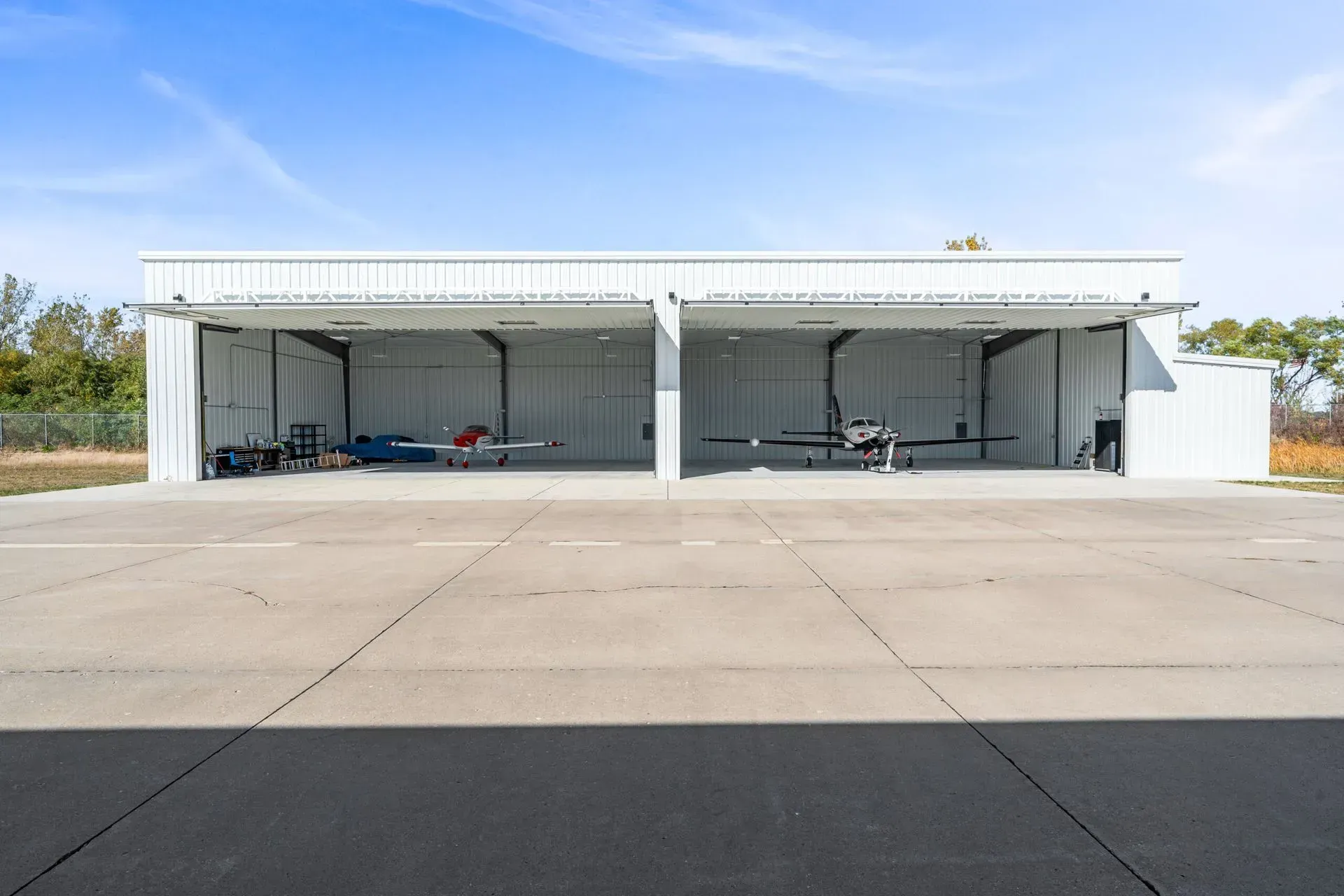 A white metal aircraft hangar with two open bays, each housing a small airplane, set against a clear blue sky.