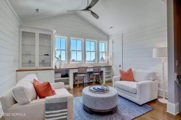 A bright, modern home office with shiplap walls, two white sofas, a central ottoman, and a built-in desk under windows.