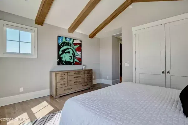 A bedroom with a dresser, pop-art Statue of Liberty painting, wooden ceiling beams, and a white bed.
