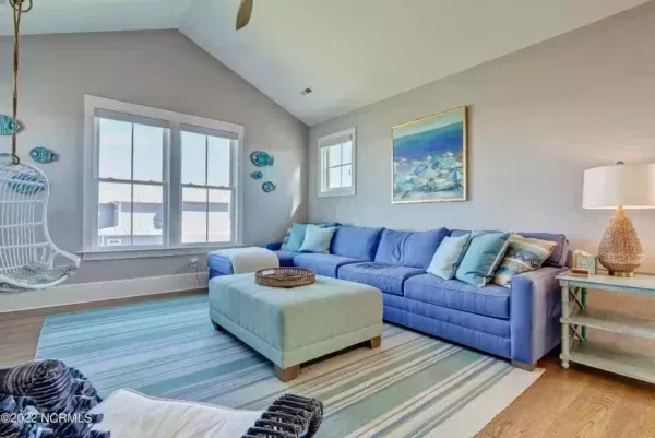 A coastal-themed living room features a blue sectional sofa, a light blue ottoman, and a hanging chair on a striped rug.