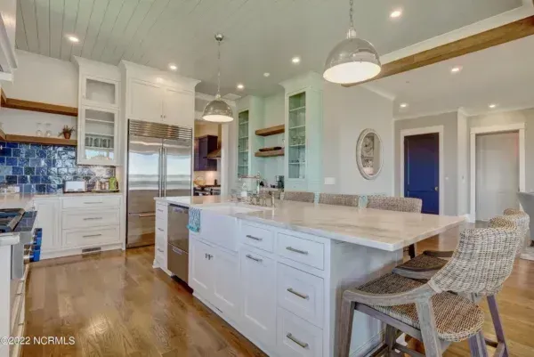 A brightly lit kitchen with a central marble island, white cabinetry, blue tiled backsplash, and woven bar stools.
