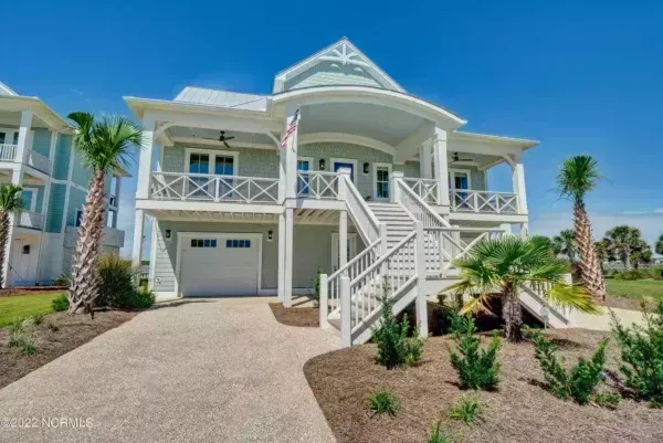 A two-story, light green beach house with a white wraparound porch, stairs to the front entrance, and a paved driveway.