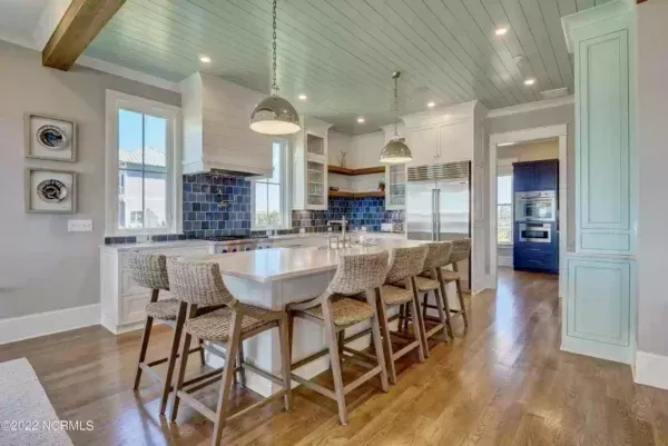 A modern kitchen with a large island, woven bar stools, light blue ceiling, and dark blue tile backsplash.