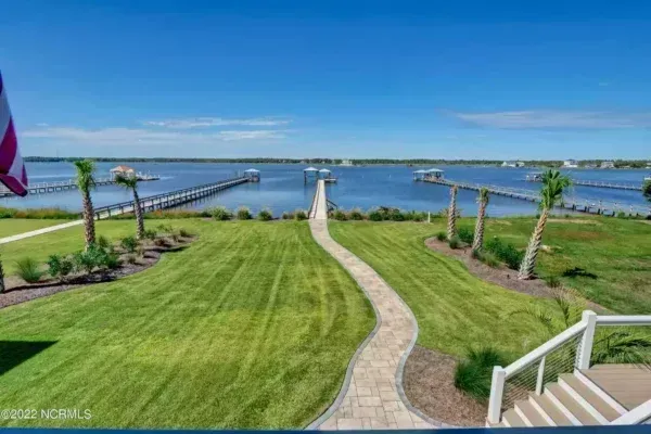 A stone walkway leads across a grassy yard toward a wooden pier extending into a calm, blue bay under a clear sky.