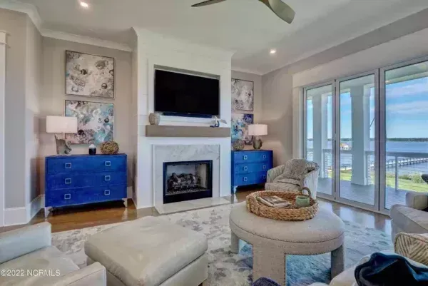 A bright living room with a fireplace, TV, blue cabinets, neutral furniture, and sliding doors overlooking the water.