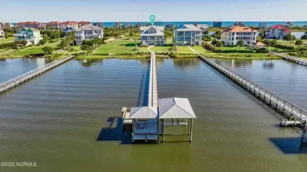 An aerial view of a waterfront house with a long wooden pier extending into the water, surrounded by other coastal homes.