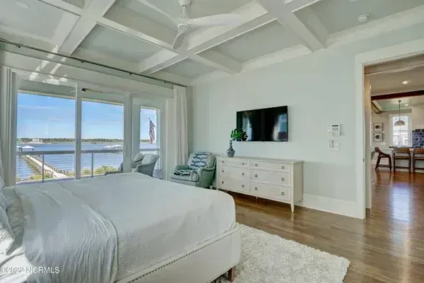 A modern bedroom with a large bed, white coffered ceiling, dresser with TV, and sliding doors overlooking the water.
