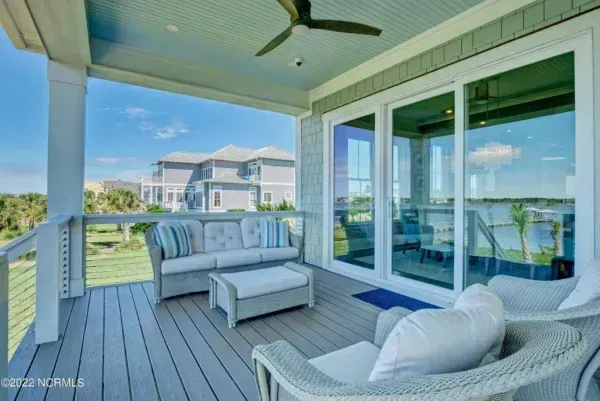 Covered deck with gray wicker outdoor furniture, a light blue ceiling fan, and a view of a coastal home.