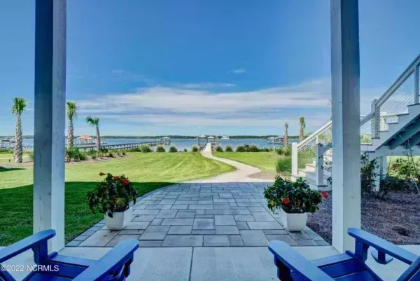 A view from a patio featuring two blue chairs, overlooking a grassy lawn, a stone path, and a waterfront with a pier.