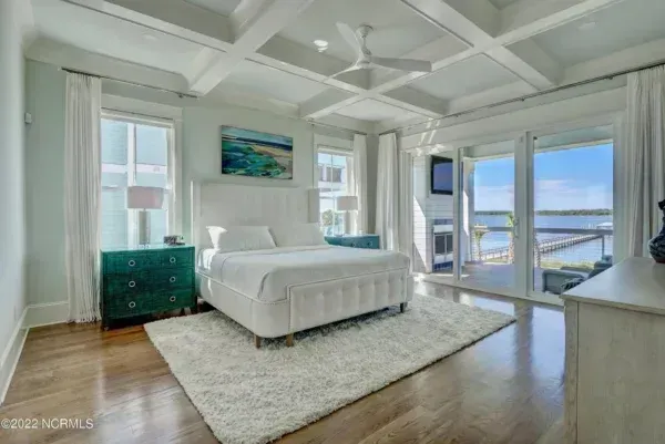A bright, airy bedroom with a white bed, teal nightstand, coffered ceiling, and a sliding glass door overlooking the water.