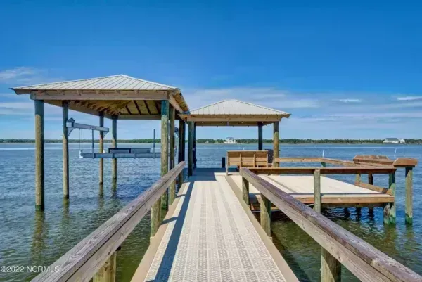 A wooden pier with two covered boat slips extends over calm water under a clear blue sky.