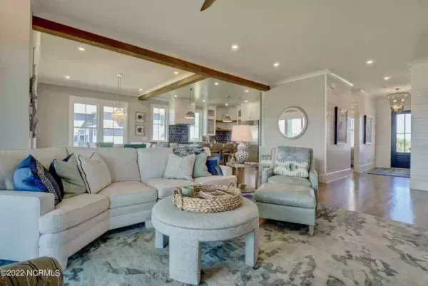A bright living room with a white sectional sofa, a round ottoman, and an armchair, opening into a kitchen area.