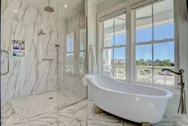 A marble-tiled walk-in shower and a freestanding white bathtub in a modern bathroom with large windows overlooking a lawn.