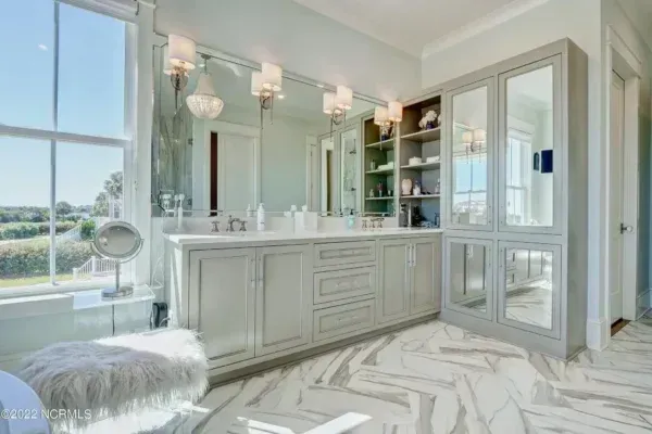 A bright, luxurious bathroom featuring gray cabinetry, a large mirror, open shelving, and chevron-patterned floor tiles.