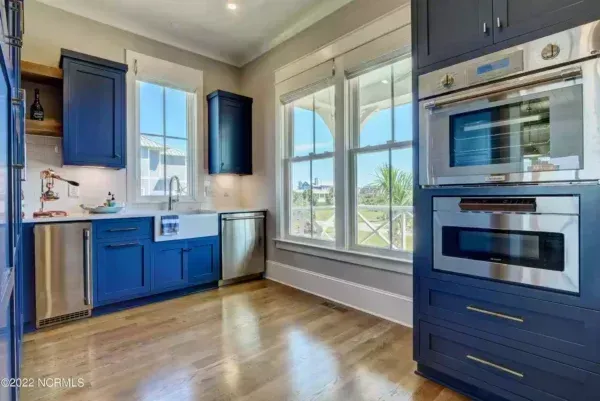 A modern kitchen with blue cabinets, hardwood floors, large windows, and built-in stainless steel double ovens.