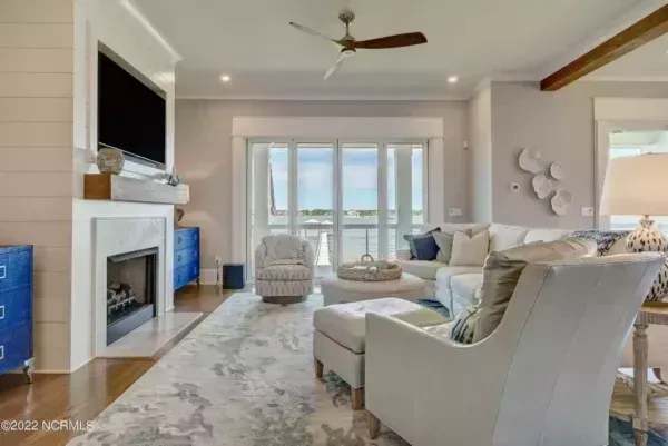 A bright living room with a fireplace, white furniture, a patterned rug, and a large sliding glass door facing the water.