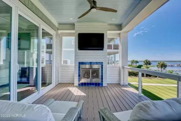 A covered outdoor porch with a fireplace, wall-mounted television, ceiling fan, and lounge chairs overlooking a bay.