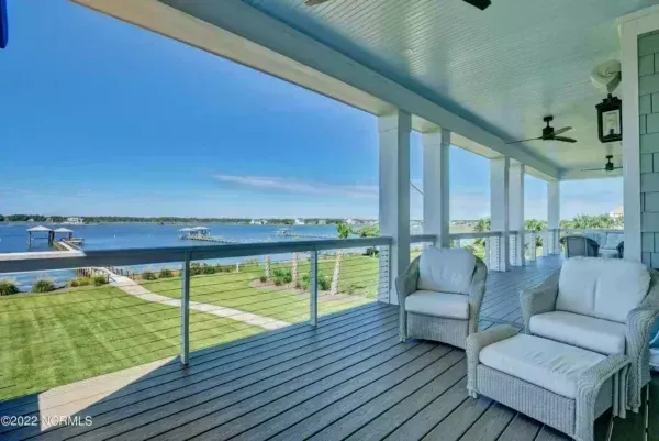 A spacious, covered wooden deck with outdoor seating, overlooking a grassy lawn and a blue body of water.