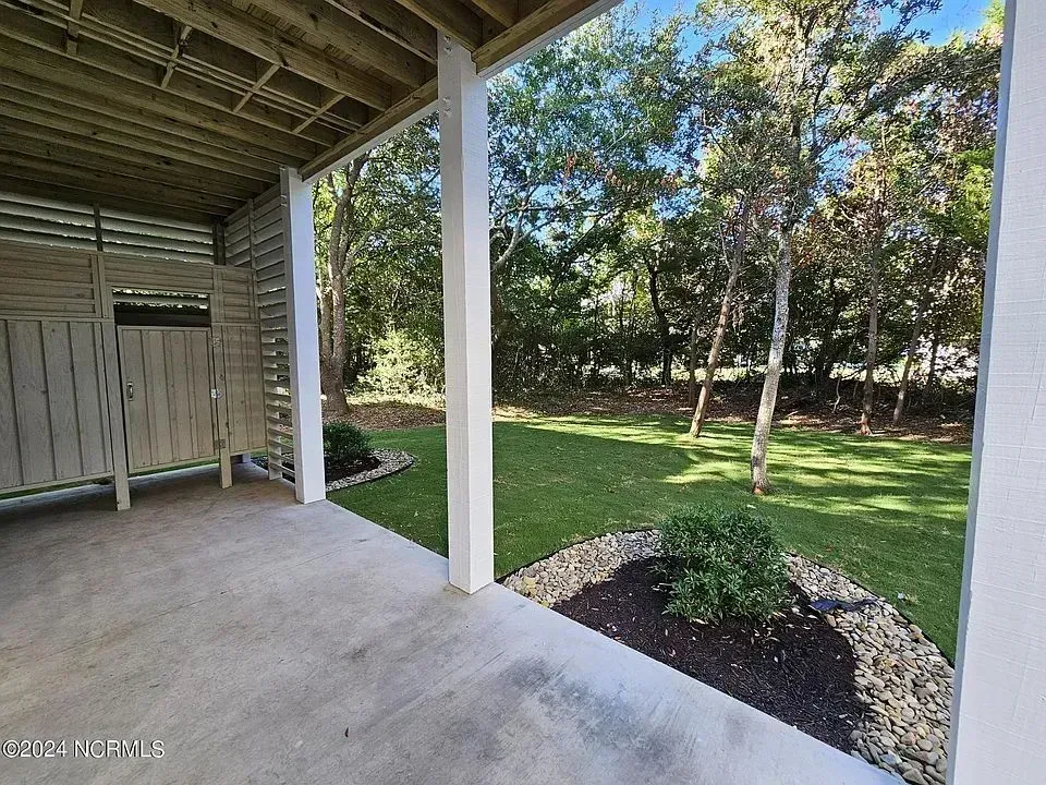 A covered concrete patio overlooks a backyard with a patch of mulch, a green lawn, and surrounding trees.