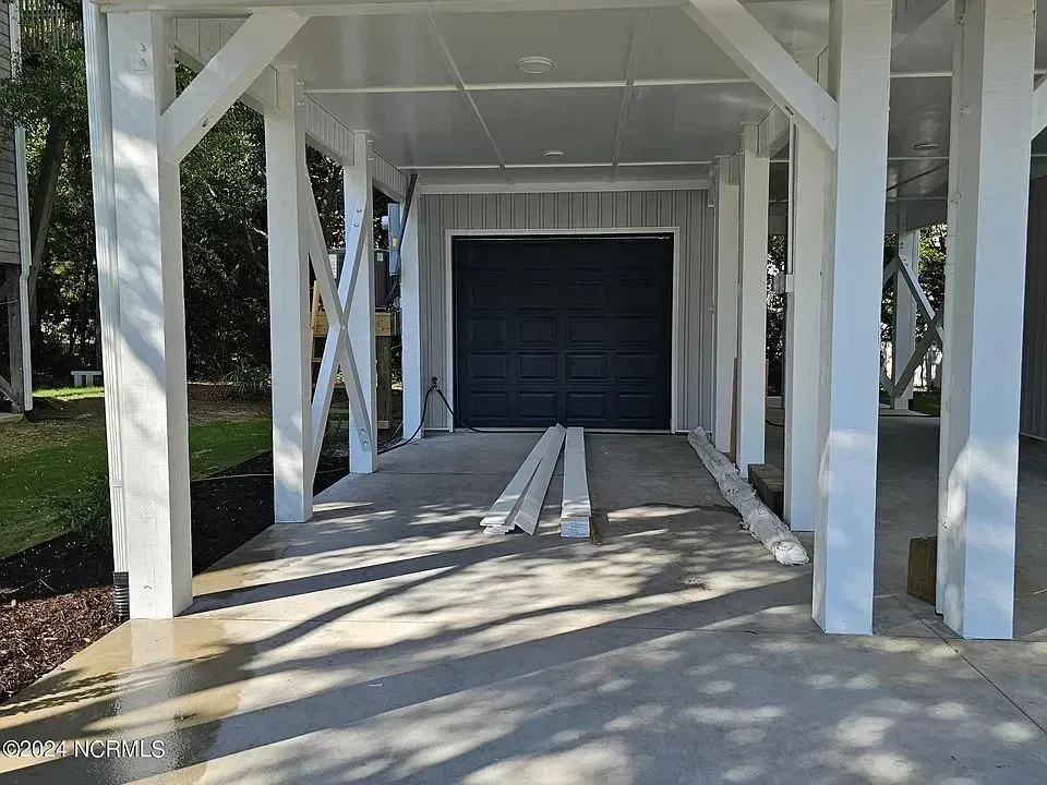 A ground-level view of a house on stilts with a dark blue garage door and white structural columns over a concrete slab.