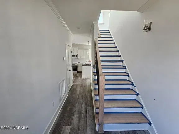 A staircase with carpeted steps and wooden banisters leads upward in a hallway with light-colored walls and wood flooring.