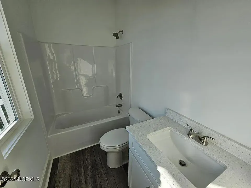 A modern, bright bathroom with a white vanity sink, toilet, and tub-shower combo against white walls and wood-look flooring.