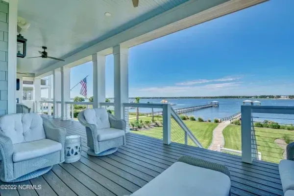 A covered wooden porch with armchairs overlooking a lawn, a pier, and a calm body of water under a clear blue sky.