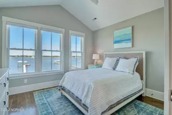 A serene bedroom with a bed, light blue area rug, and windows overlooking a calm body of water with a dock.