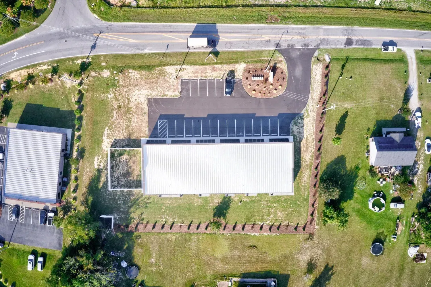 Aerial view of a commercial building with a metal roof, parking lot, and landscaping, situated near a road.